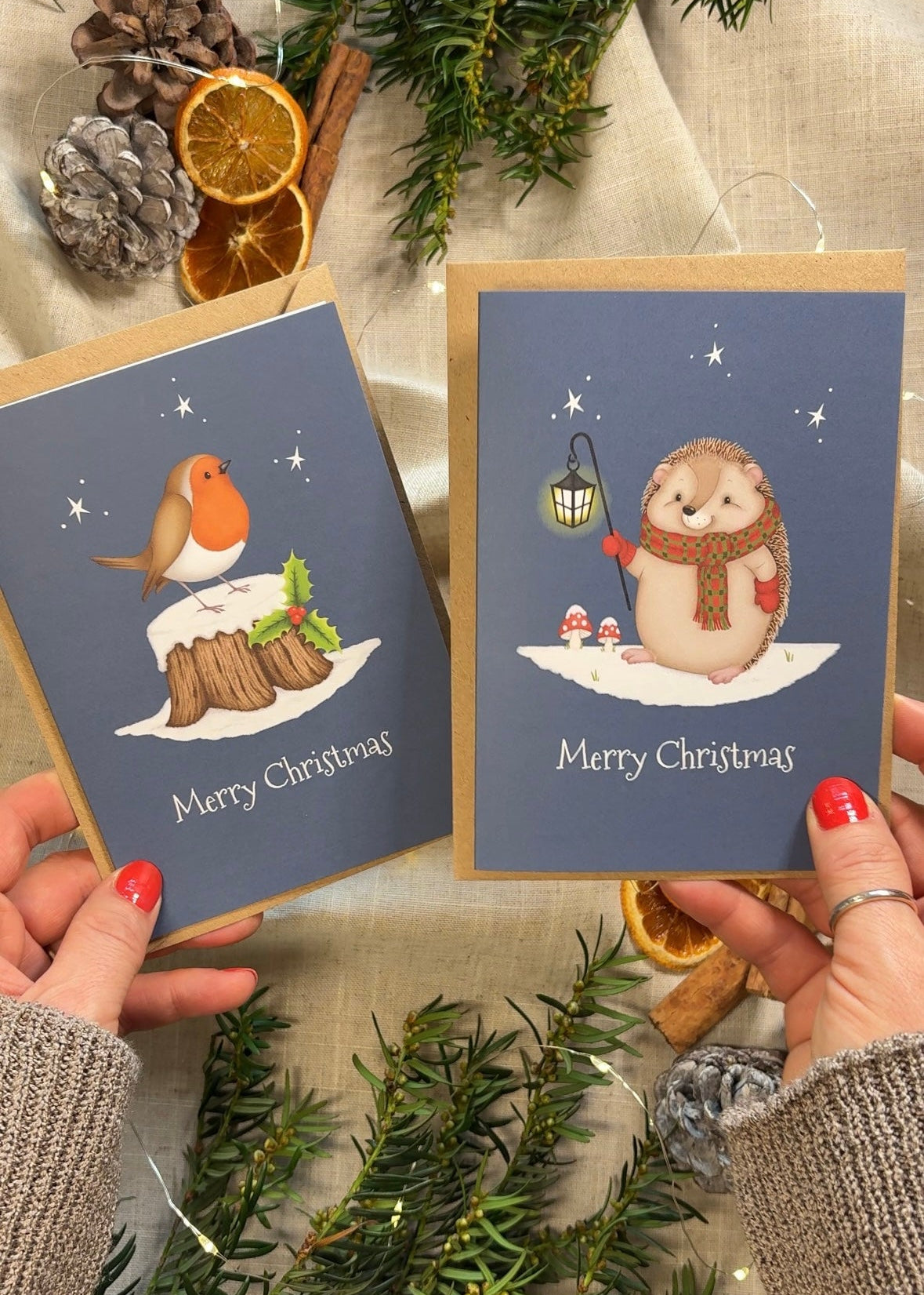 Two Christmas cards with winter-themed illustrations held by hands, surrounded by festive decorations.